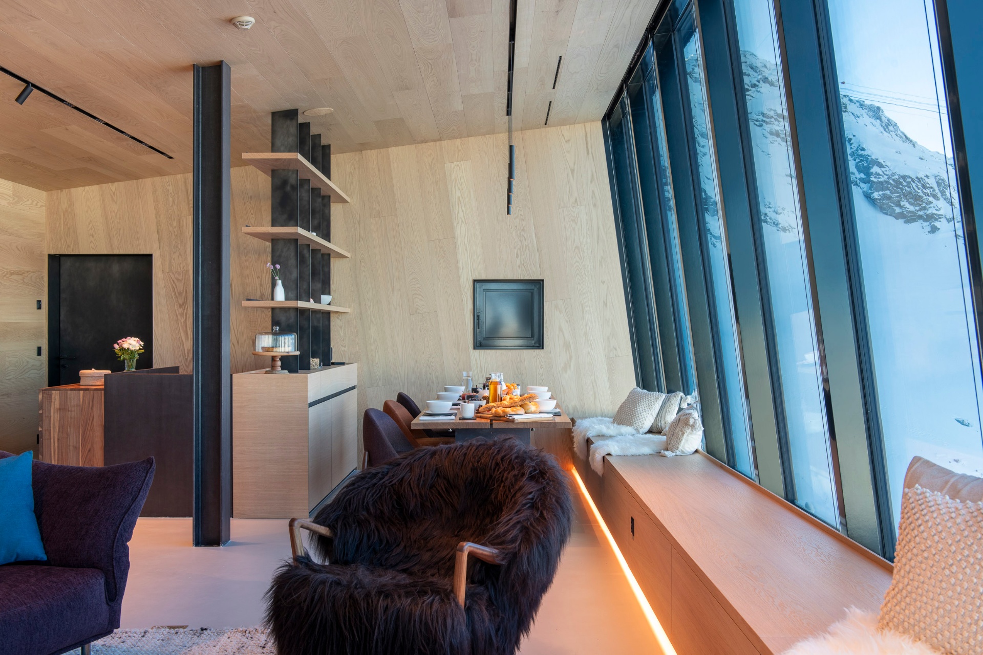 Elegant private dining table set for guests inside Cabane Tortin with floor-to-ceiling windows overlooking snow-covered Alpine peaks at sunset